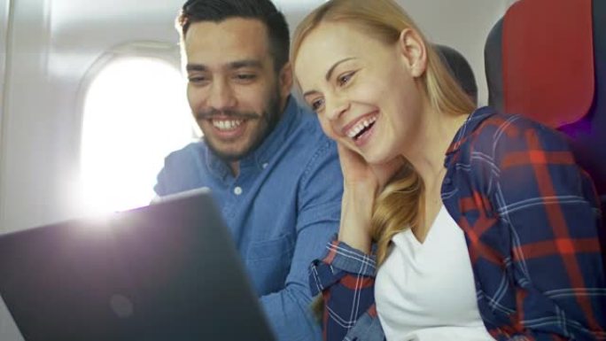 在商用飞机上，漂亮的年轻金发女郎和英俊的西班牙裔男性在笔记本电脑上看电影并大笑。阳光透过飞机的窗户照