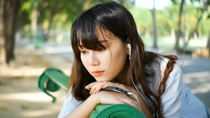 美丽的年轻潮人悲伤的亚洲女人用智能手机戴着耳机在情感上听音乐，坐在公园的长椅上，在夏天日落的阳光下。