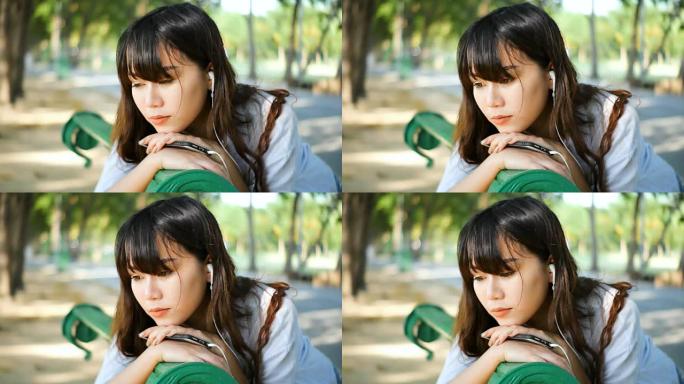 美丽的年轻潮人悲伤的亚洲女人用智能手机戴着耳机在情感上听音乐，坐在公园的长椅上，在夏天日落的阳光下。