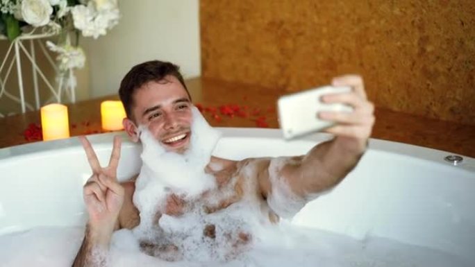 帅哥正在用智能手机在浴缸里自拍，胡须上有肥皂泡沫。他笑着做手势，摆出姿势，玩得开心。