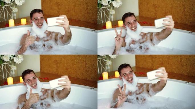 帅哥正在用智能手机在浴缸里自拍，胡须上有肥皂泡沫。他笑着做手势，摆出姿势，玩得开心。