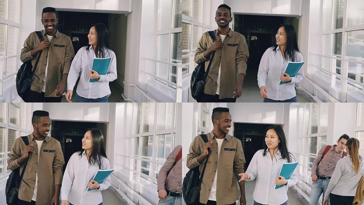 年轻的有吸引力的非洲男性和亚洲女学生正在走廊上移动，讨论一些生动活泼的事情，并示意另一对站在高窗附近