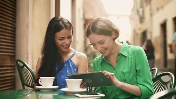 两个穿着浅色夏装的迷人年轻女性在街头咖啡店放松和交谈。一个人在平板电脑上分享一些令人兴奋的东西。