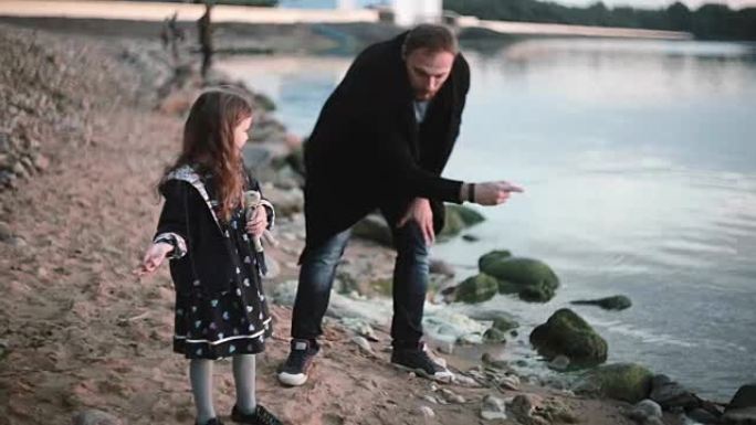 父女在岸上，小女孩向水中扔石头。男人向女孩展示如何玩石头跳绳