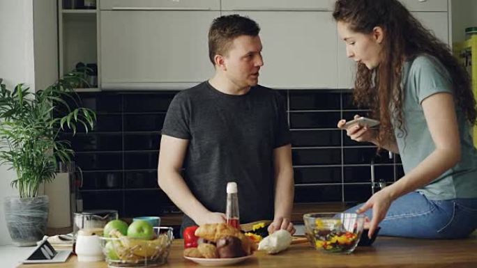 迷人的年轻夫妇在家里的厨房里做饭，愉快地聊天。男人切蔬菜做沙拉，他的女朋友在社交媒体的智能手机相机上