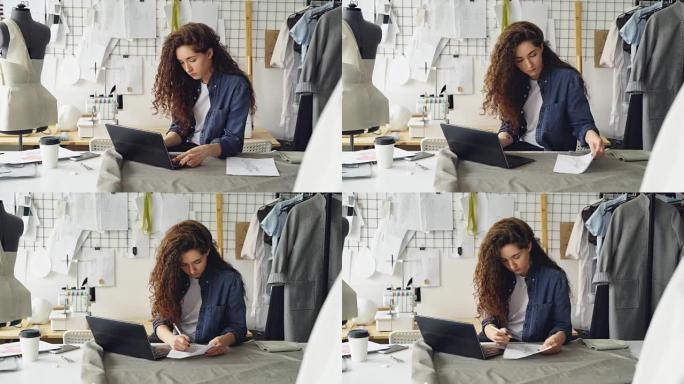 自雇的女裁缝正在现代照明车间的餐桌旁与笔记本电脑一起工作。女人在打字，看碎石，然后画服装素描。