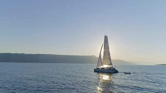 一艘帆船双体船的空中长镜头，帆船在平静的海面上航行，太阳升起，沿海丘陵可见。人们在甲板上放松。