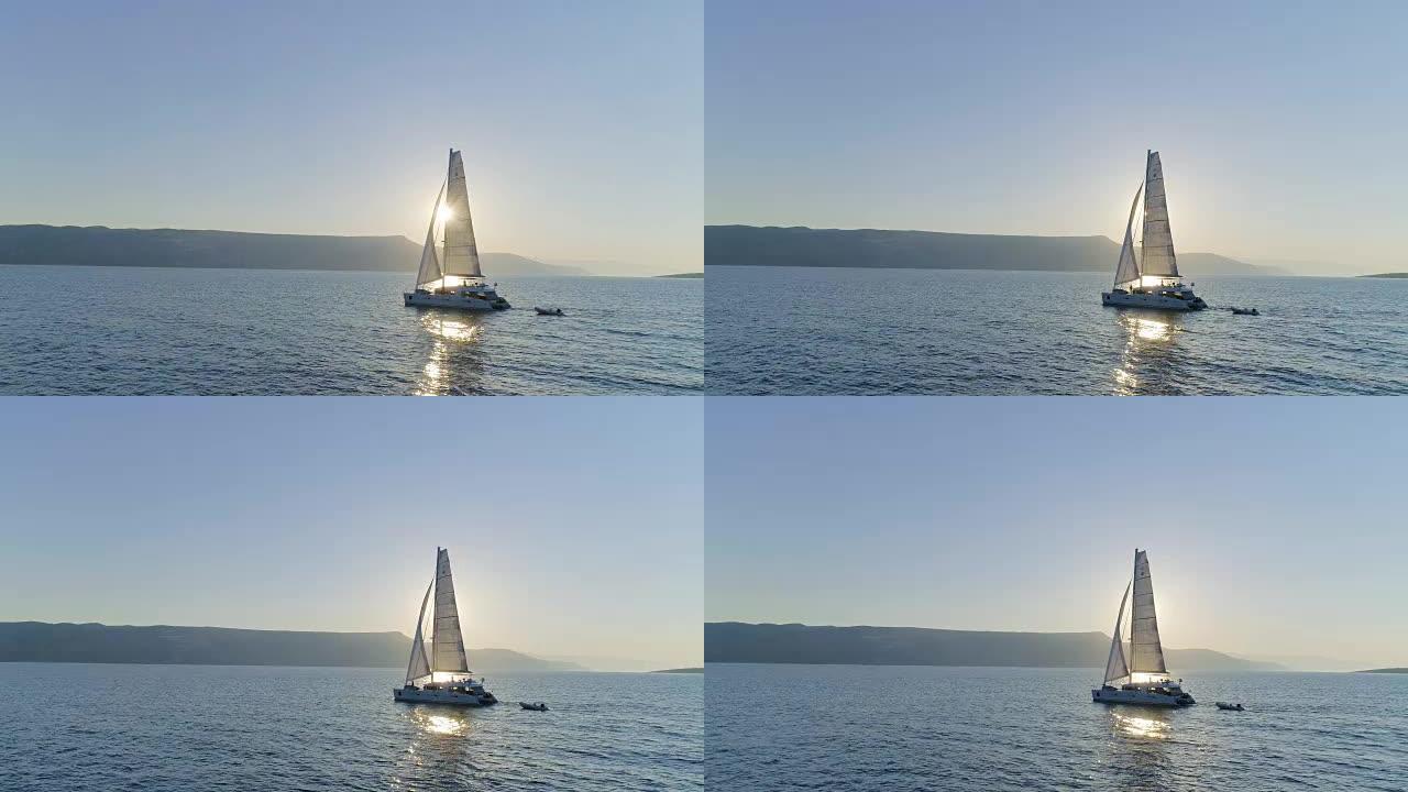 一艘帆船双体船的空中长镜头，帆船在平静的海面上航行，太阳升起，沿海丘陵可见。人们在甲板上放松。