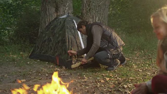 男人正在森林里搭帐篷，而两个女孩正坐在火炉旁。