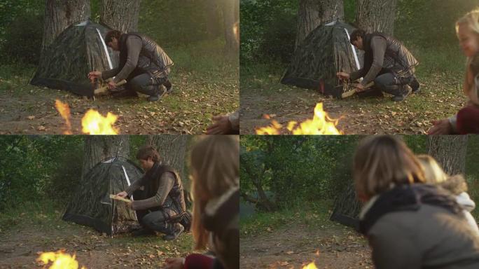 男人正在森林里搭帐篷，而两个女孩正坐在火炉旁。