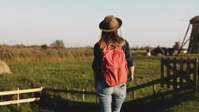 休闲女孩跳过风车农场附近的篱笆。戴着帽子的长发和红色背包的女人在日落时回家。4K