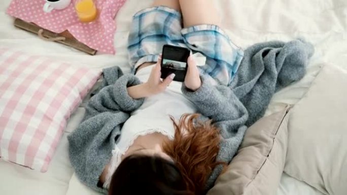 年轻漂亮的女人躺在床上，拿着手机。女孩在智能手机上观看照片并喝果汁
