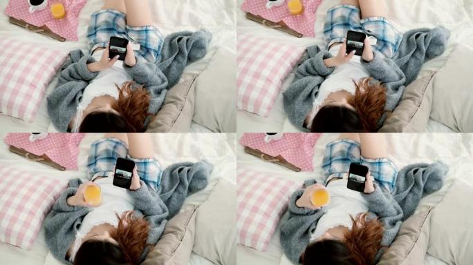 年轻漂亮的女人躺在床上，拿着手机。女孩在智能手机上观看照片并喝果汁