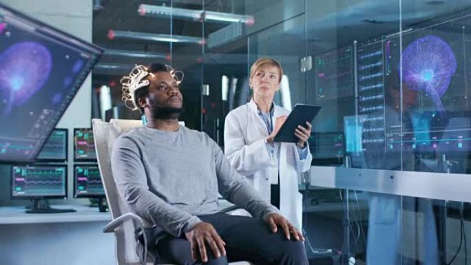 戴着脑电波扫描耳机的男人坐在椅子上，而科学家则用平板电脑监督过程。在现代的大脑研究实验室监视器显示脑