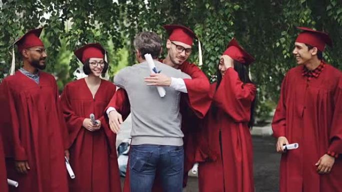 骄傲的大学老师的慢动作拥抱着他的学生并握手，即将毕业的学生站在毕业证书的队伍中大笑。