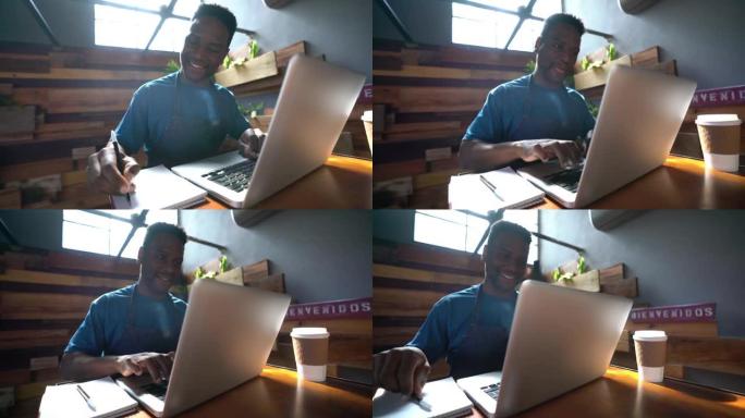 一家咖啡馆的黑人企业主在笔记本电脑上工作，在记事本上做笔记，看起来很开心