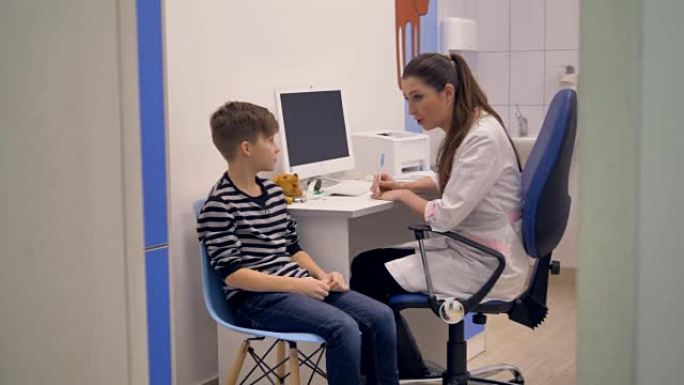 一名儿科医生在她的桌子上和一个学龄男孩交谈。