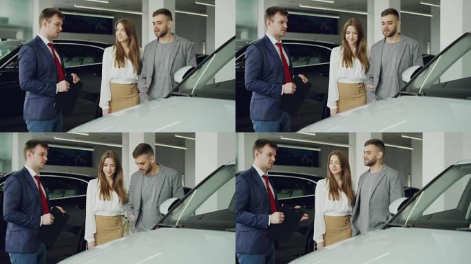 友好的汽车推销员正在与自信的年轻人交谈，向他讲述新的车型，而漂亮女人正站在丈夫旁边，抱着他的手臂微笑