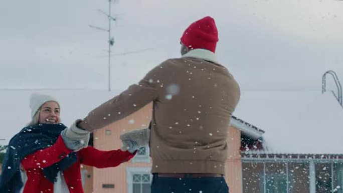 年轻美丽的夫妇在飘落的雪中跳舞和旋转。快乐的男人和女人在他们田园诗般的房子的院子里玩得开心。享受冬天