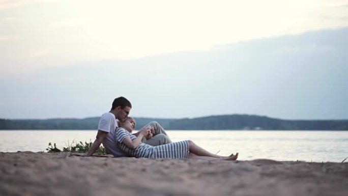 美丽的年轻夫妇坐在河边，使用智能手机。男人和女人在沙滩上休息