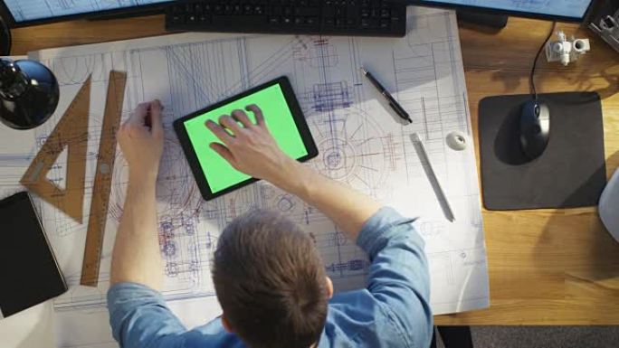 建筑工程师使用蓝图的俯视图，使用带有绿色屏幕的平板电脑，与蓝图进行比较。他的桌子上摆满了有用的物品和
