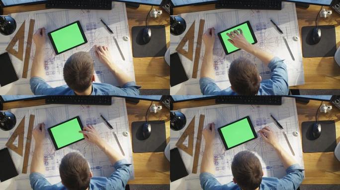 建筑工程师使用蓝图的俯视图，使用带有绿色屏幕的平板电脑，与蓝图进行比较。他的桌子上摆满了有用的物品和