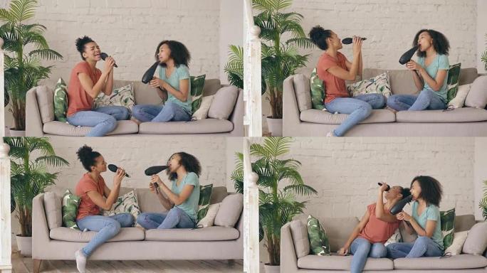 混合种族年轻有趣的女孩坐在沙发上用吹风机和梳子跳舞唱歌。姐妹们在客厅享受休闲的乐趣