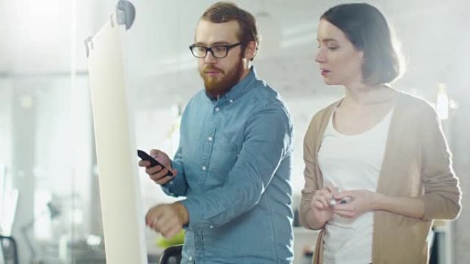 年轻的男人和一个女人在白板上讨论工作过程。男人拿着智能手机。女人在白板上画画。
