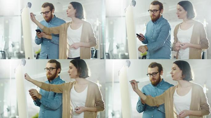 年轻的男人和一个女人在白板上讨论工作过程。男人拿着智能手机。女人在白板上画画。