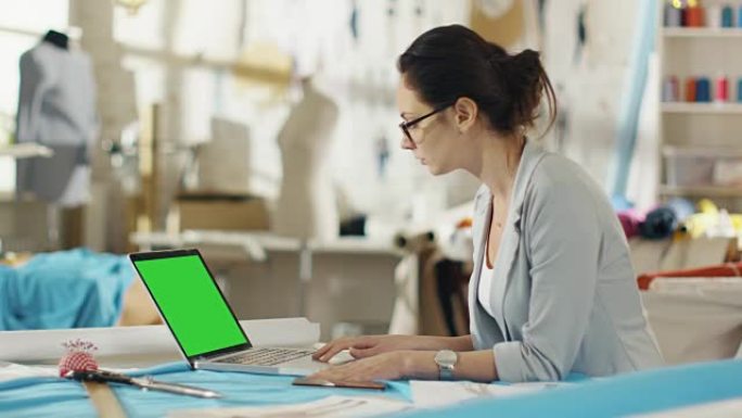 美丽的女性时装设计师坐在她的办公桌前，在绿屏笔记本电脑上工作。她的工作室阳光明媚，色彩丰富的面料和人