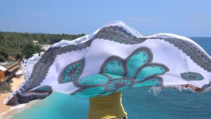 慢镜头女孩拿着飘动的围巾在巴厘岛的沙滩和蓝色的大海上