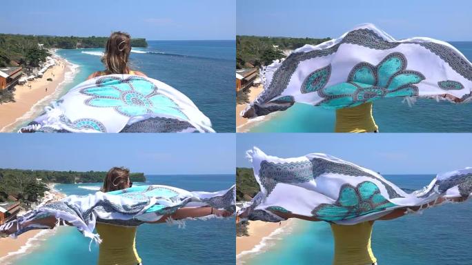 慢镜头女孩拿着飘动的围巾在巴厘岛的沙滩和蓝色的大海上