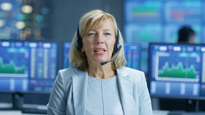 首席销售人员代表与耳机交谈。在工作人员的背后，屏幕上显示了股市行情数字和图表。