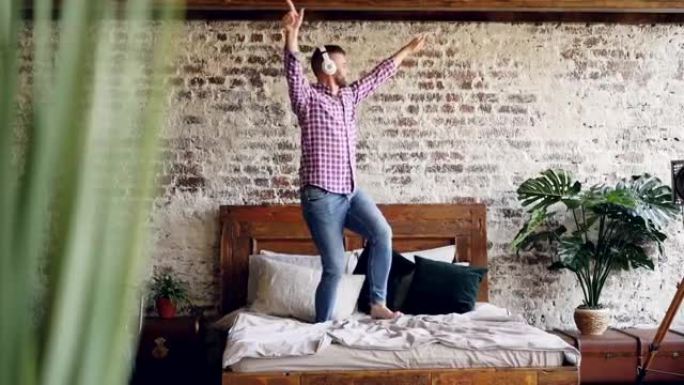 年轻的大胡子男人在双人床上跳舞，戴着耳机听音乐。现代阁楼风格的公寓和植物是可见的。