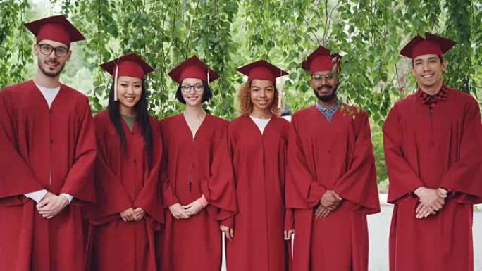 站在户外的多种族毕业学生的肖像穿着红色礼服和灰泥板，微笑着看着相机。