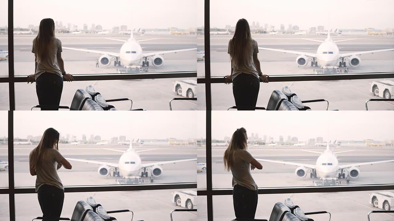 年轻的欧洲女商人站在机场候机厅的窗户上看，看着并使用智能手表