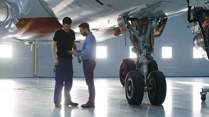 在机库，飞机维修工程师向飞机技术员展示平板电脑上的技术数据。他们站在干净的新飞机旁边。