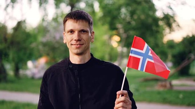 快乐的挪威学生手持官方国旗，看着相机微笑的慢动作肖像。美丽的绿色公园是背景。