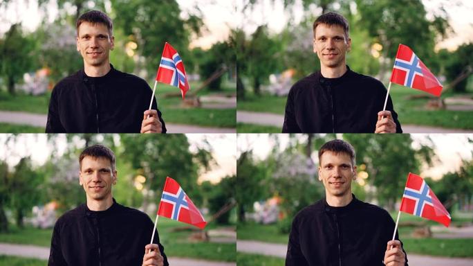 快乐的挪威学生手持官方国旗，看着相机微笑的慢动作肖像。美丽的绿色公园是背景。