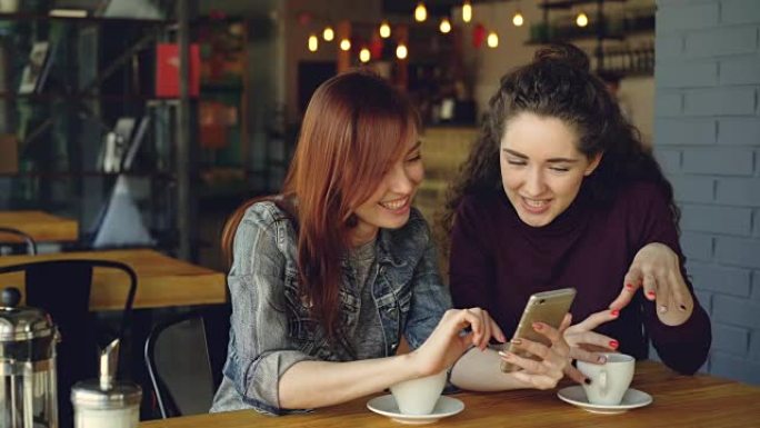 两个年轻漂亮的女友在咖啡馆里使用智能手机，在情感上大笑和交谈。社交媒体、会友与现代科技理念。