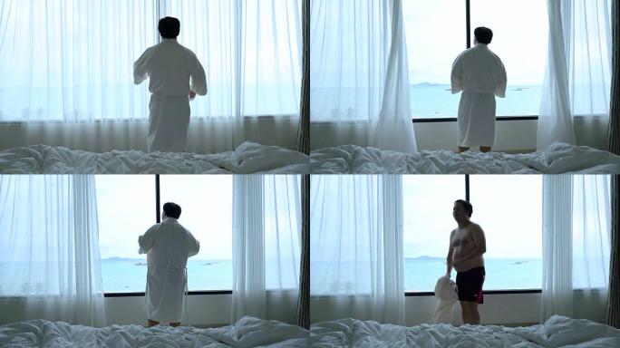 4k镜头亚洲男子步行打开窗户上的窗帘并在早晨在豪华酒店卧室醒来时脱下浴袍的场景，生活方式和休闲概念
