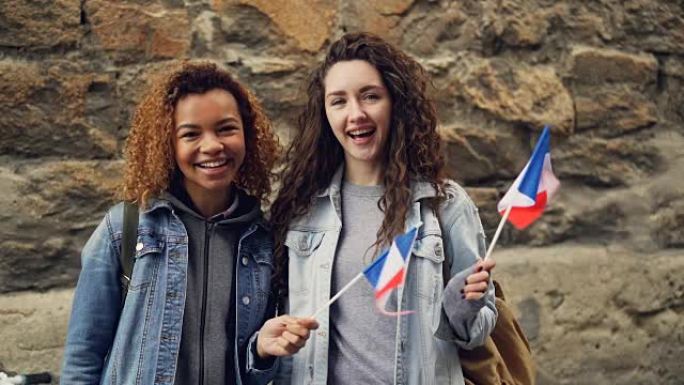 两个笑着的女学生挥舞着法国国旗，看着站在砖墙上的相机的慢动作肖像。友谊、旅游和幸福的人的概念。