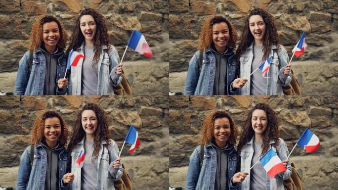 两个笑着的女学生挥舞着法国国旗，看着站在砖墙上的相机的慢动作肖像。友谊、旅游和幸福的人的概念。
