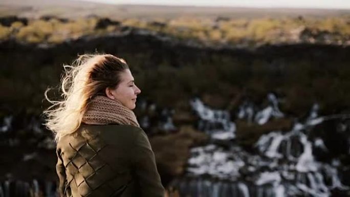 年轻的金发女子站在悬崖上，看着冰岛的巴纳福斯瀑布，抚摸着头发