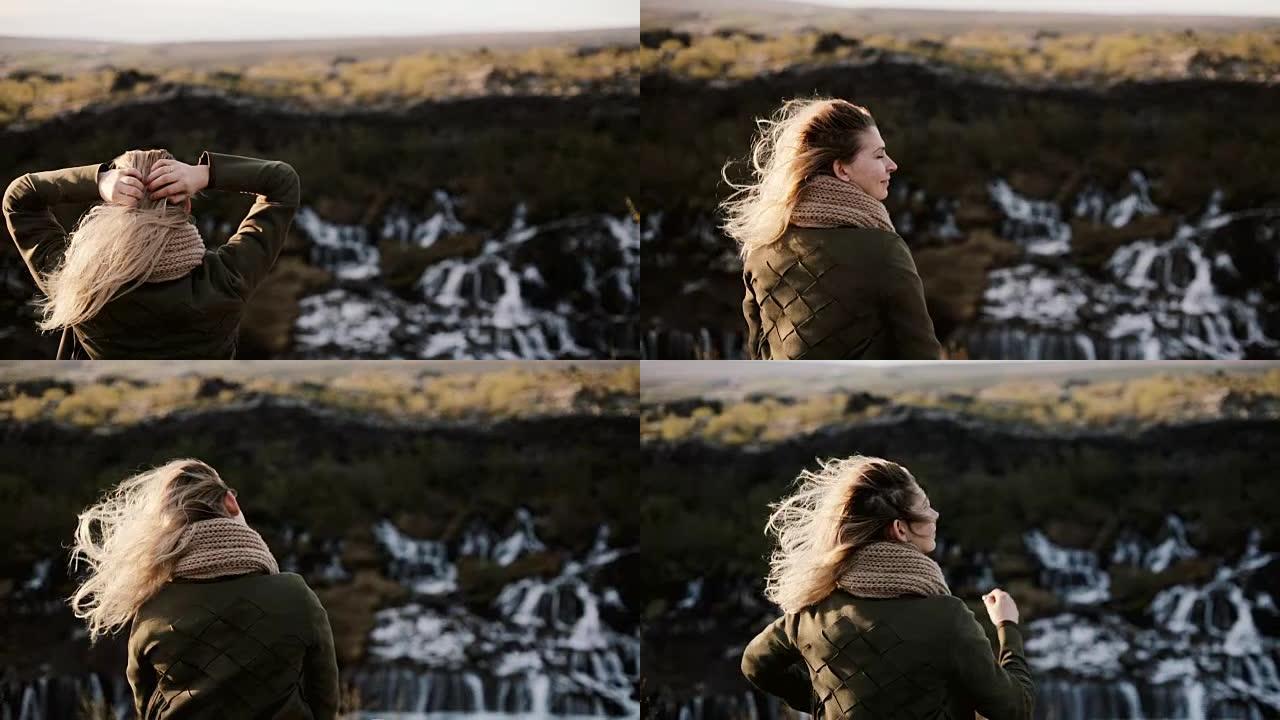 年轻的金发女子站在悬崖上，看着冰岛的巴纳福斯瀑布，抚摸着头发