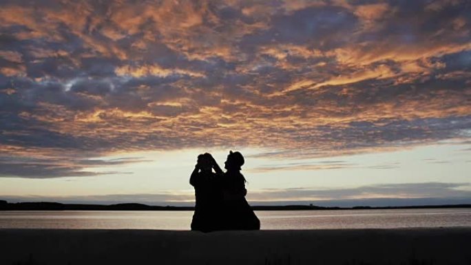 一对时尚的恋爱年轻夫妇的剪影坐在海边的长廊上，背景是美丽的日落天空