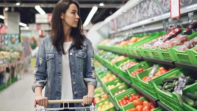 漂亮的年轻女子正沿着水果和蔬菜行，推着购物车，微笑着看着有机食品。健康的生活方式和超市理念。
