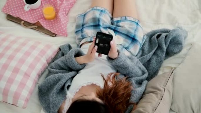 年轻的黑发女人躺在床上。早餐时使用智能手机的漂亮女孩。女性观看旅行中的照片