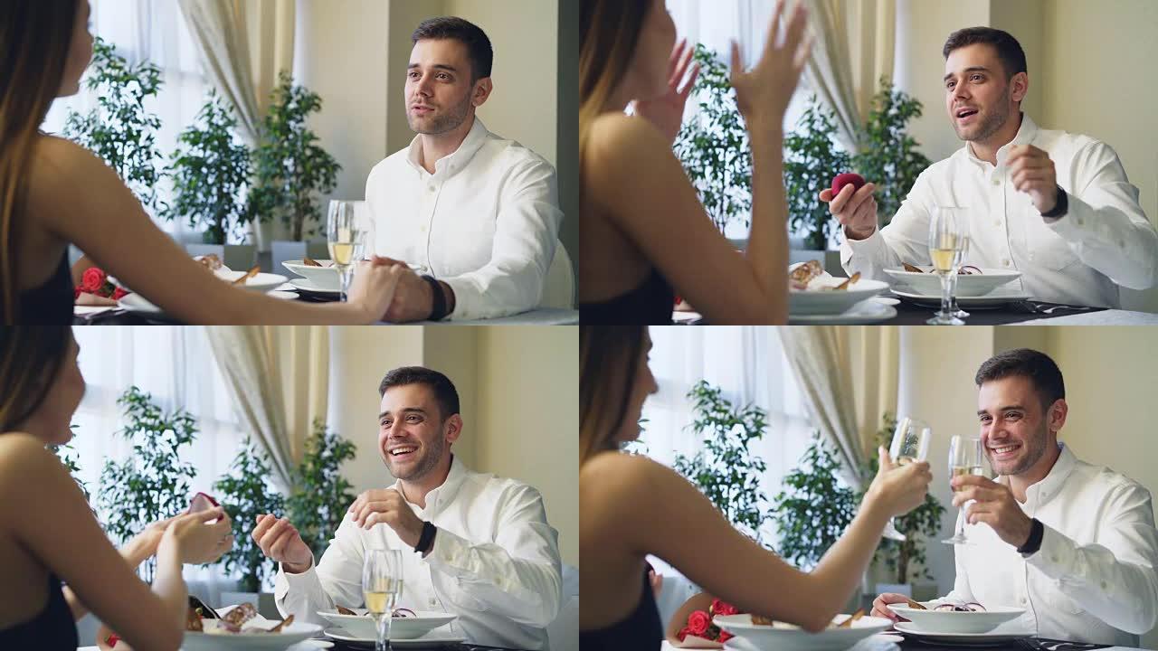 穿着白衬衫的快乐爱心男人向惊讶的美丽女友求婚，然后在餐厅浪漫约会时给她订婚戒指。