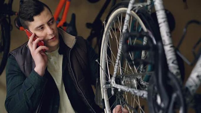严肃的年轻人自行车修理工正在用手机说话时检查和转动车轮。自行车维护、沟通和专业理念。